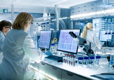 Female and Male Scientists Working on their Computers In Big Modern Laboratory. Various Shelves with Beakers, Chemicals and Different Technical Equipment is Visible.